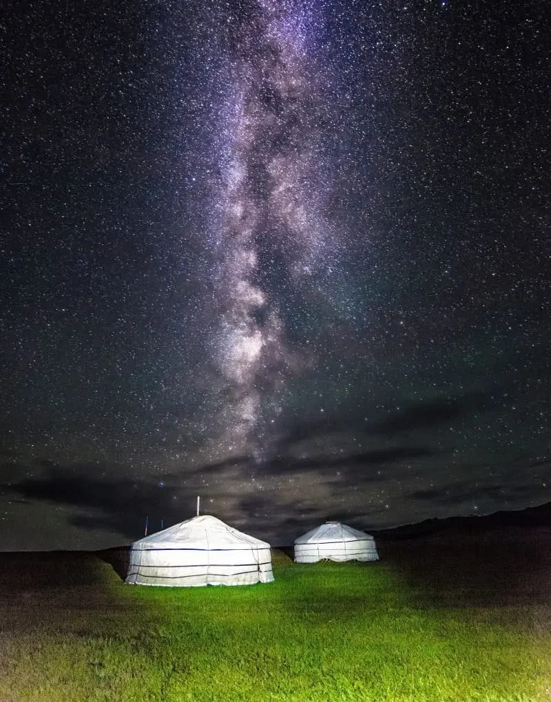 "Two traditional Mongolian ger yurts illuminated under a starry Milky Way sky in a serene night setting."